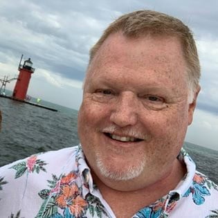 Selfie of a smiling man in a floral shirt with a red lighthouse in the background