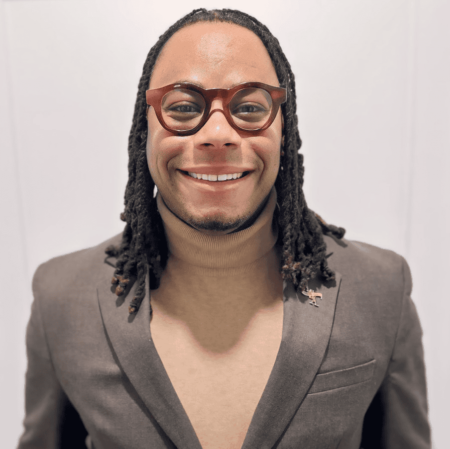 Smiling person with glasses and long dreadlocks wearing a gray blazer and tan turtleneck