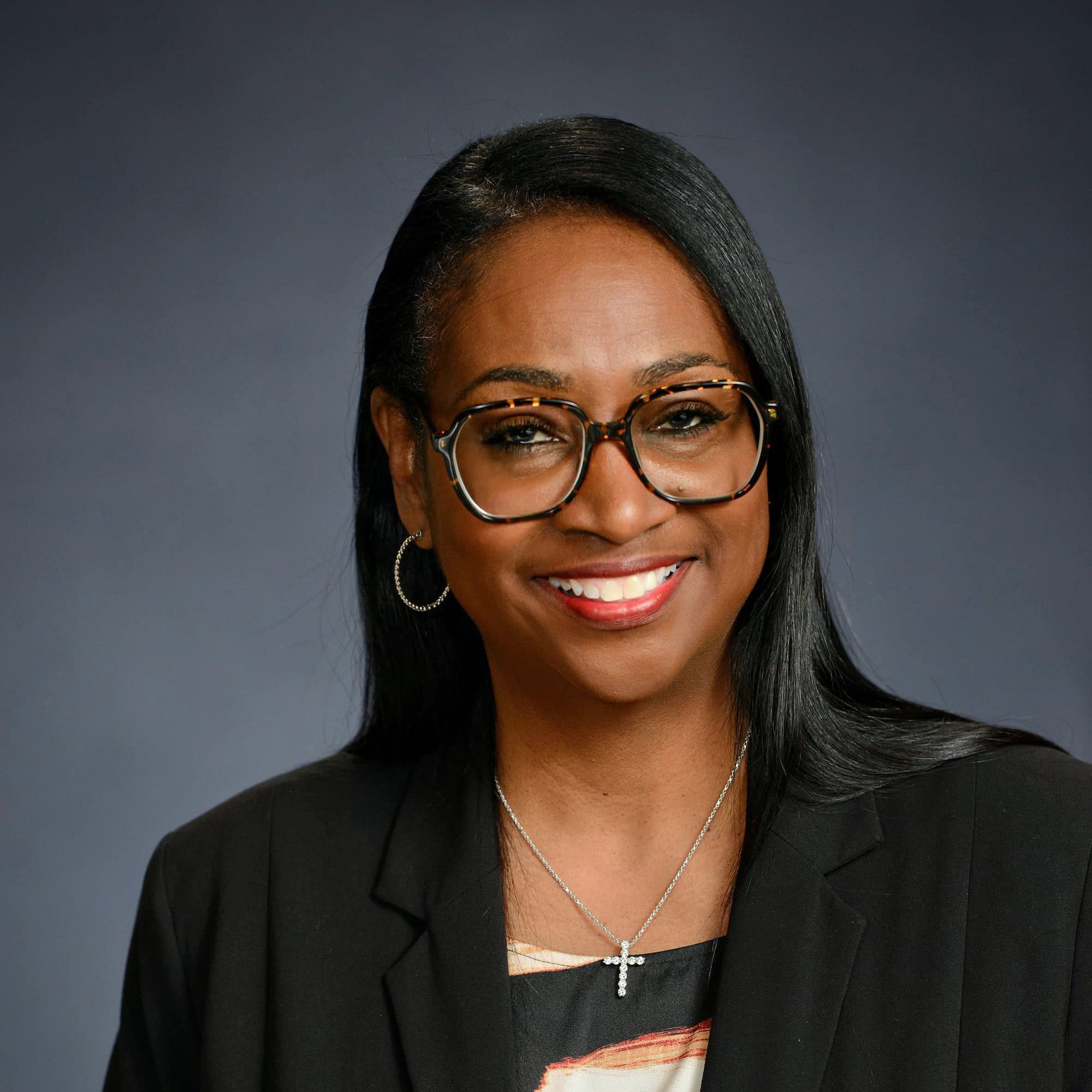 Portrait of a smiling woman wearing glasses, a black blazer, and a cross necklace against a gray background