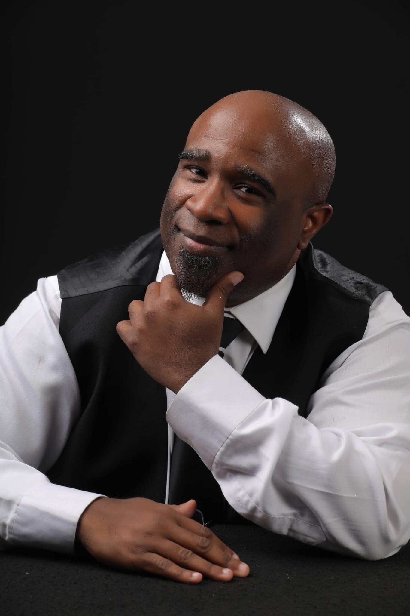 Portrait of a man in a white shirt, black vest, and tie, resting his chin on his hand against a black background