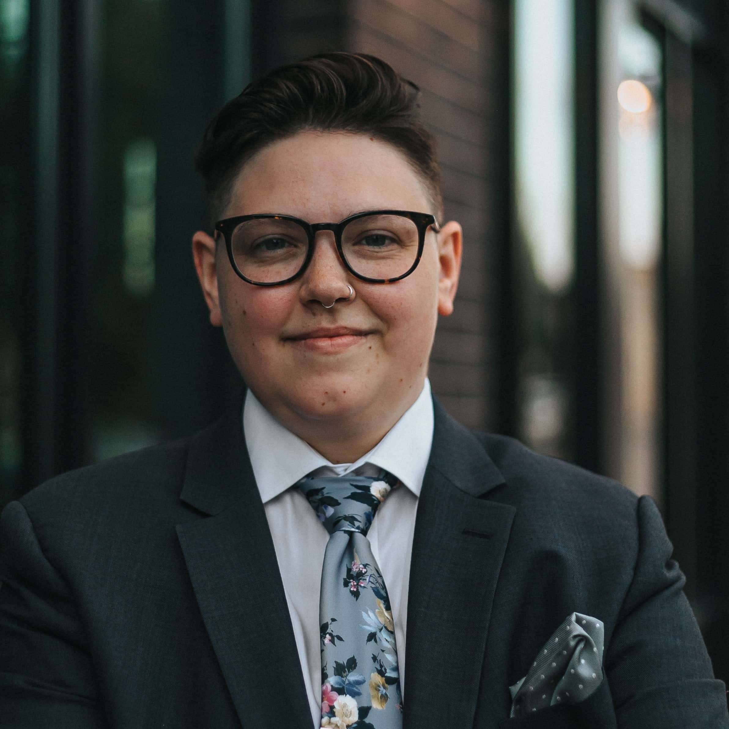 headshot of an unidentified person wearing glasses and a grey suit with a floral tie