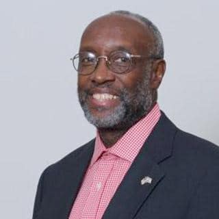 Portrait of a smiling man wearing glasses, a dark suit, and a pink checkered shirt