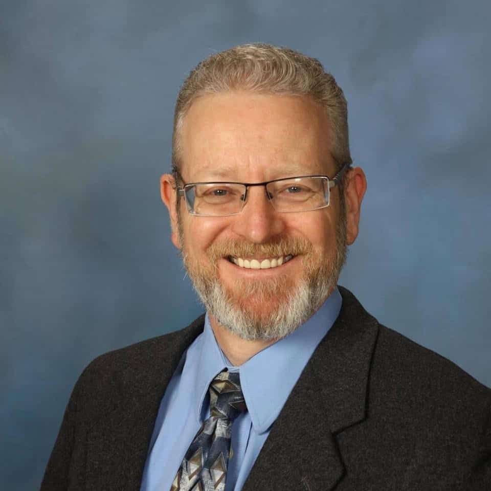 Portrait of a smiling man wearing glasses, a suit, and a patterned tie
