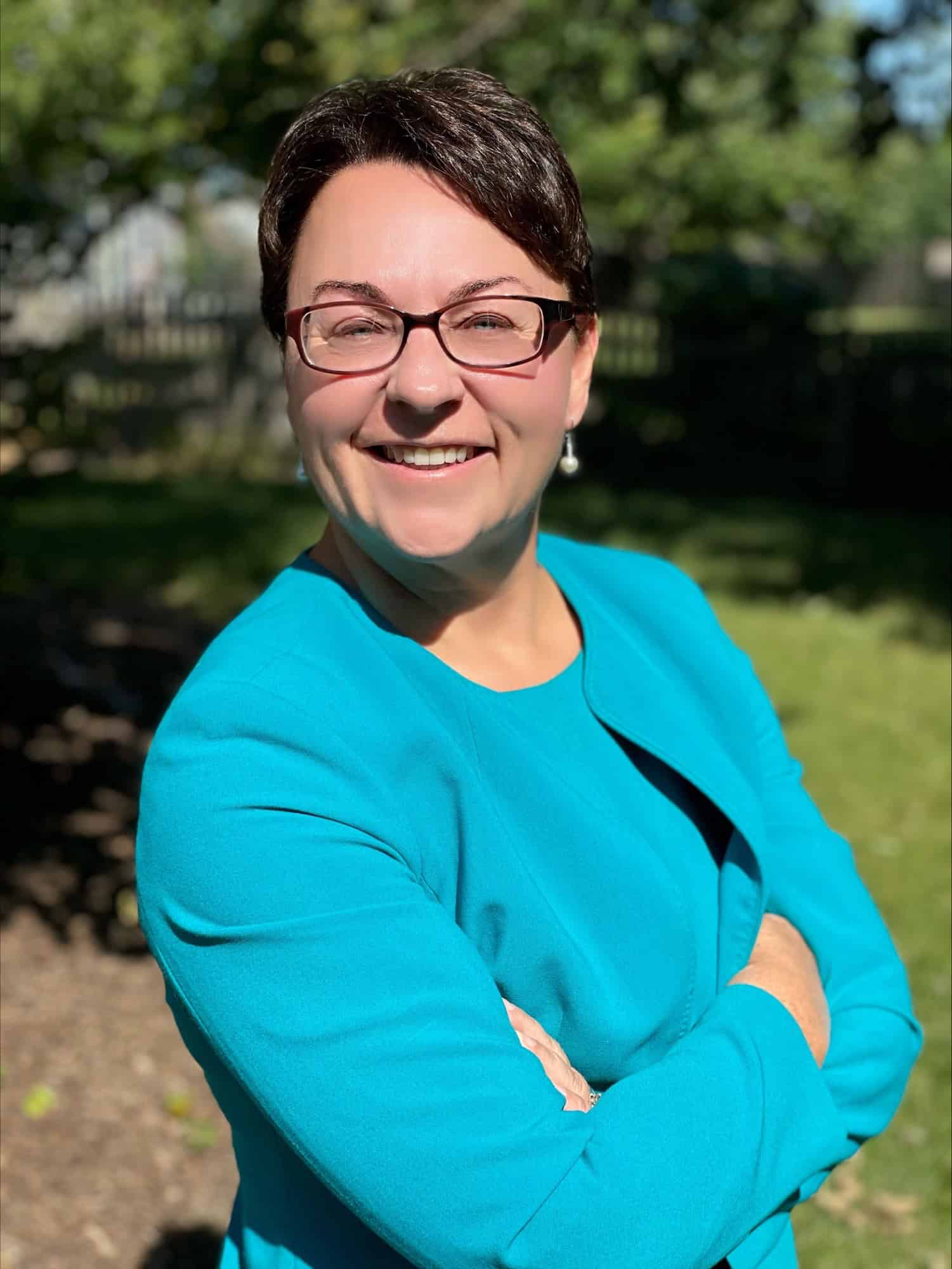 Smiling woman in a turquoise jacket and glasses standing outdoors with her arms crossed