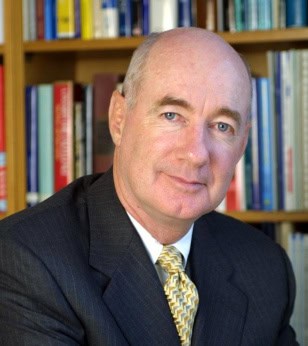 Portrait of a bald man in a suit and tie in front of bookshelves