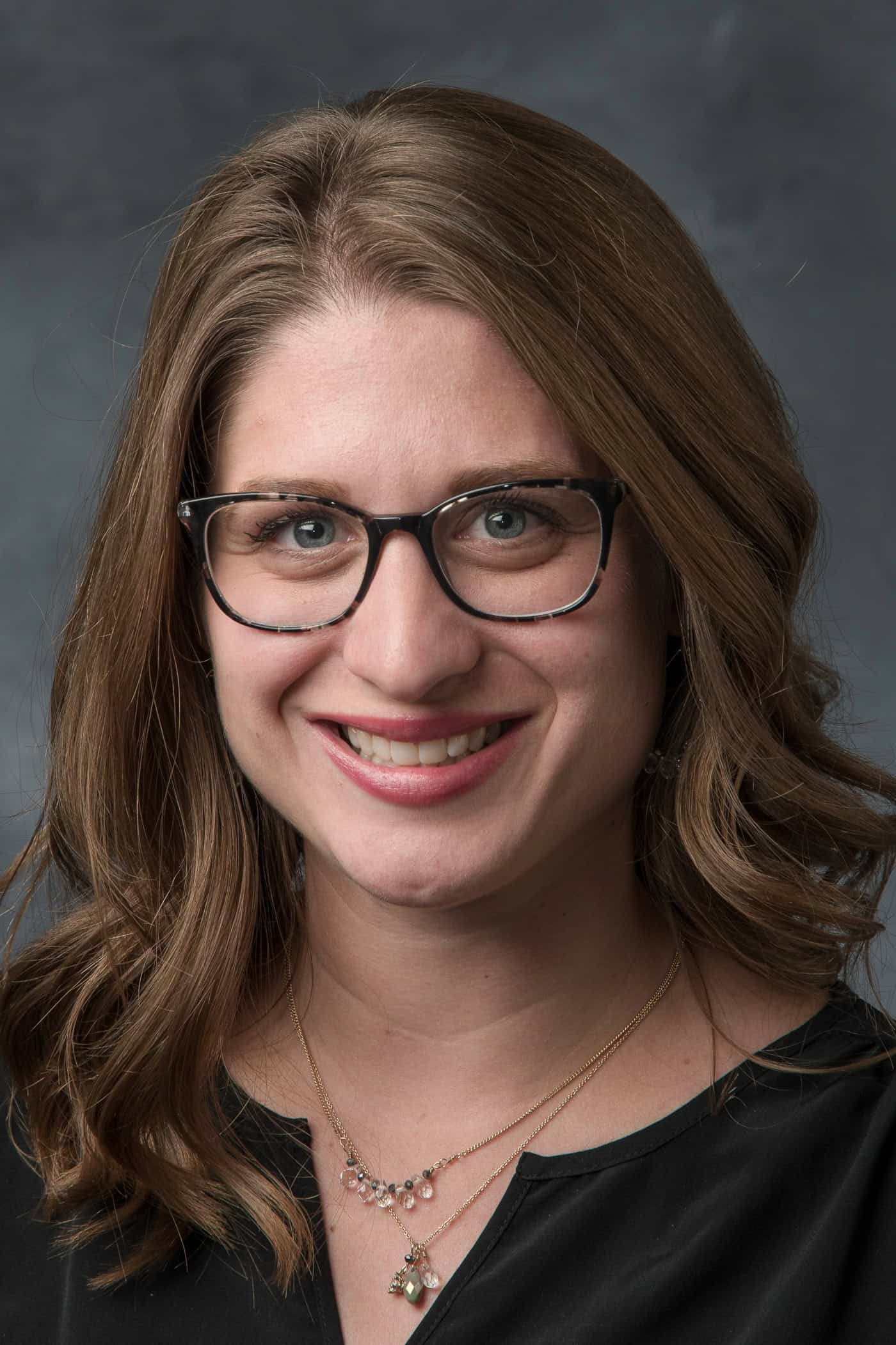 Portrait of a smiling woman wearing glasses and a necklace against a gray background