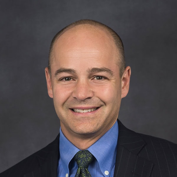 Portrait of a smiling bald man in a suit, blue shirt, and tie against a dark background