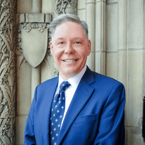 Portrait of a smiling man in a blue suit and patterned tie standing in front of an ornate stone wall
