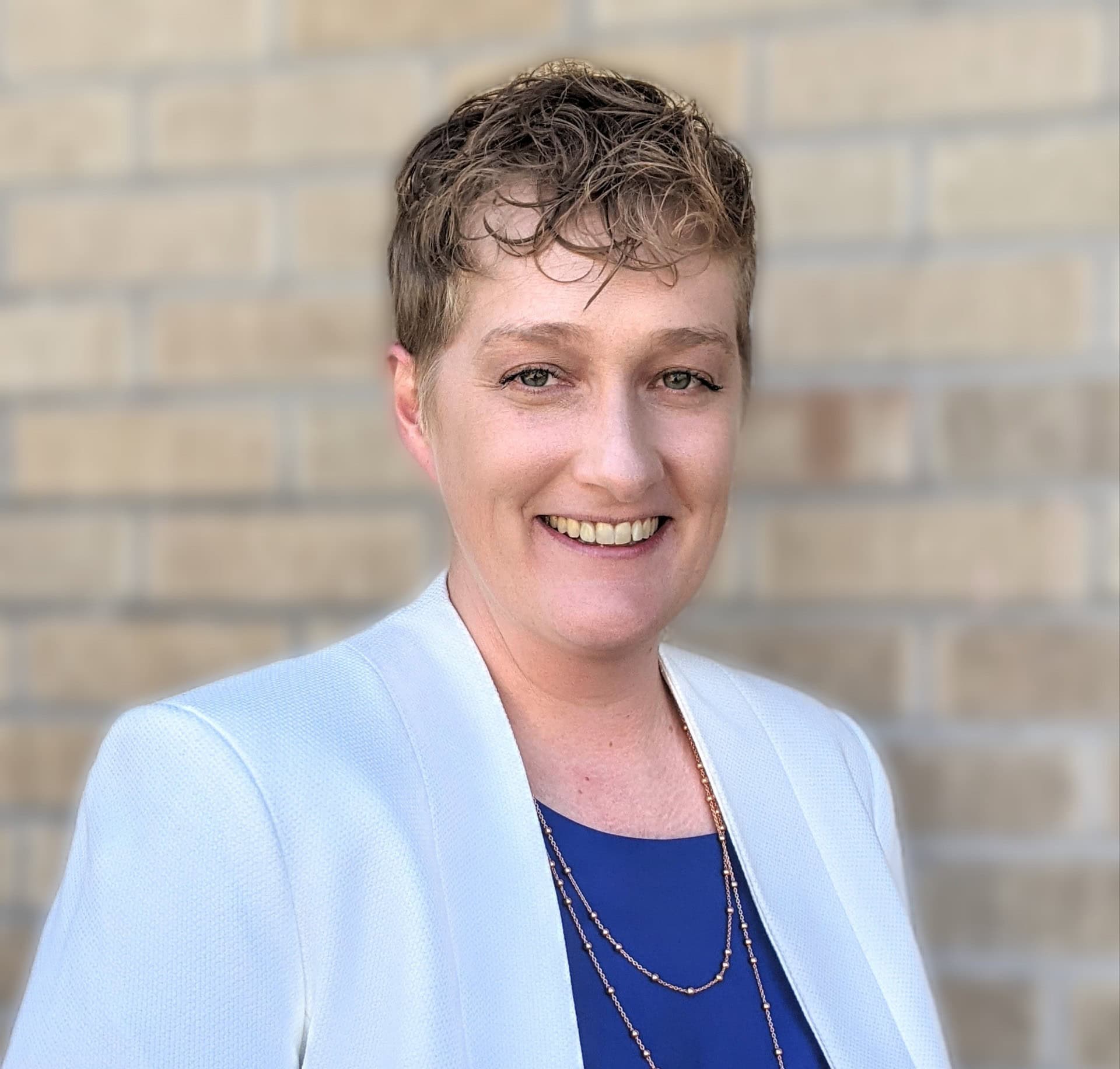 Portrait of a smiling person in a light blazer and blue top against a brick wall