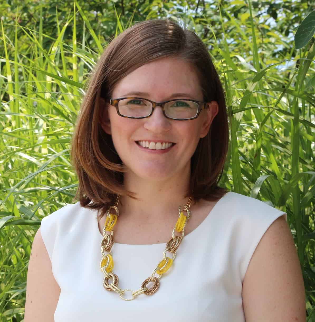 Portrait of a smiling woman wearing glasses and a white top, standing in front of tall green plants