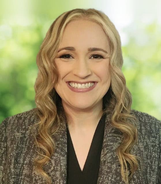 Smiling woman with long wavy blonde hair in a dark blazer against a blurred green background