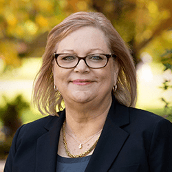 Portrait of a smiling woman wearing glasses and a dark blazer outdoors with a blurred autumn background