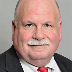 Headshot of a bald man with a white mustache in a suit and red tie