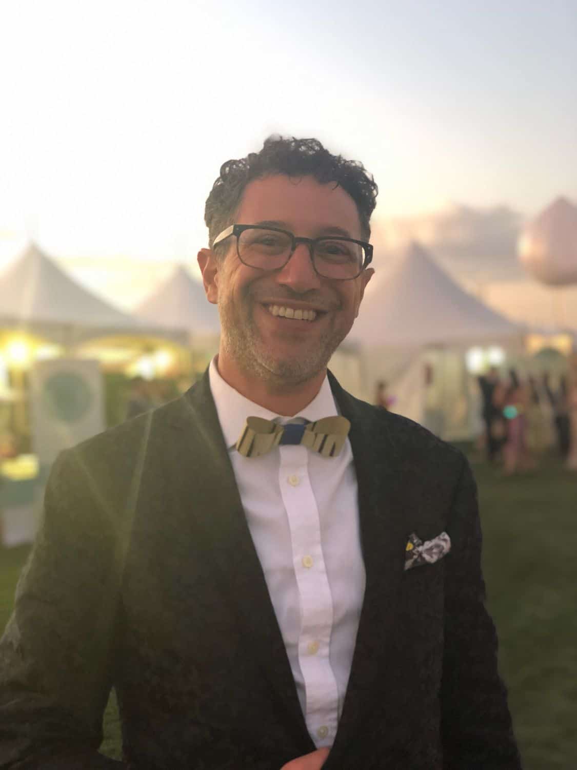 Smiling man in a suit and bow tie at an outdoor event at sunset