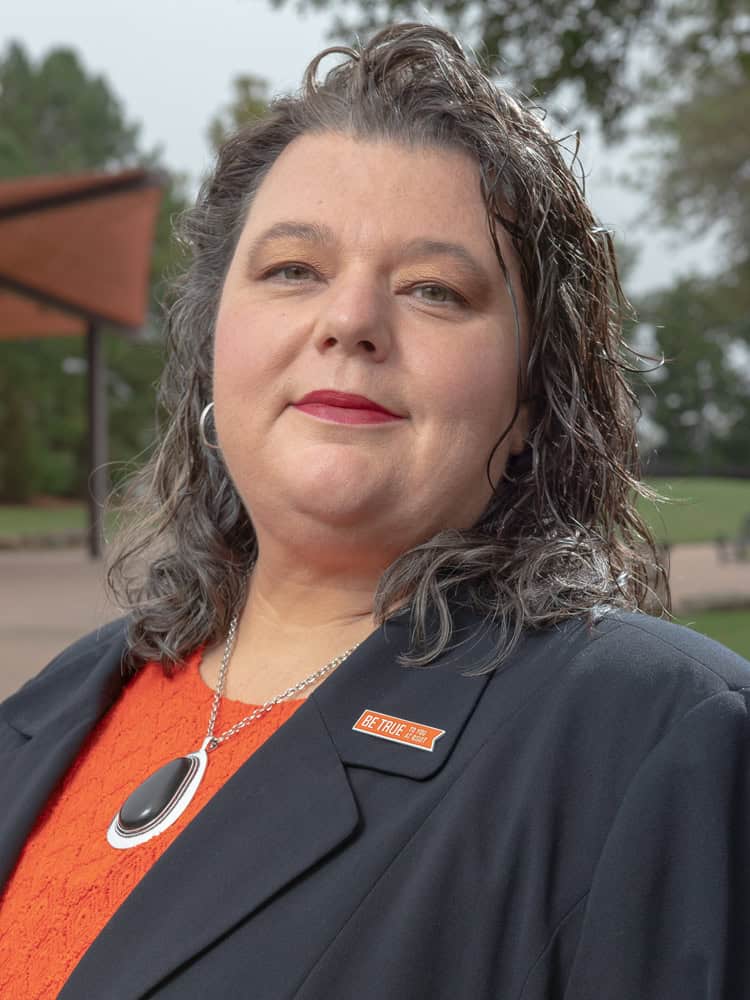 Portrait of a woman outdoors wearing a black blazer, orange top, and a “Be True” pin