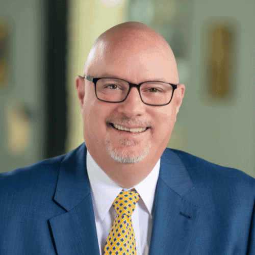 Portrait of a smiling bald man wearing glasses, a blue suit, and a yellow patterned tie