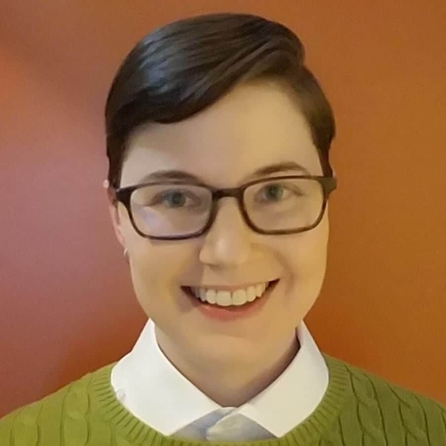 Smiling person with short dark hair and glasses wearing a green sweater over a white collared shirt