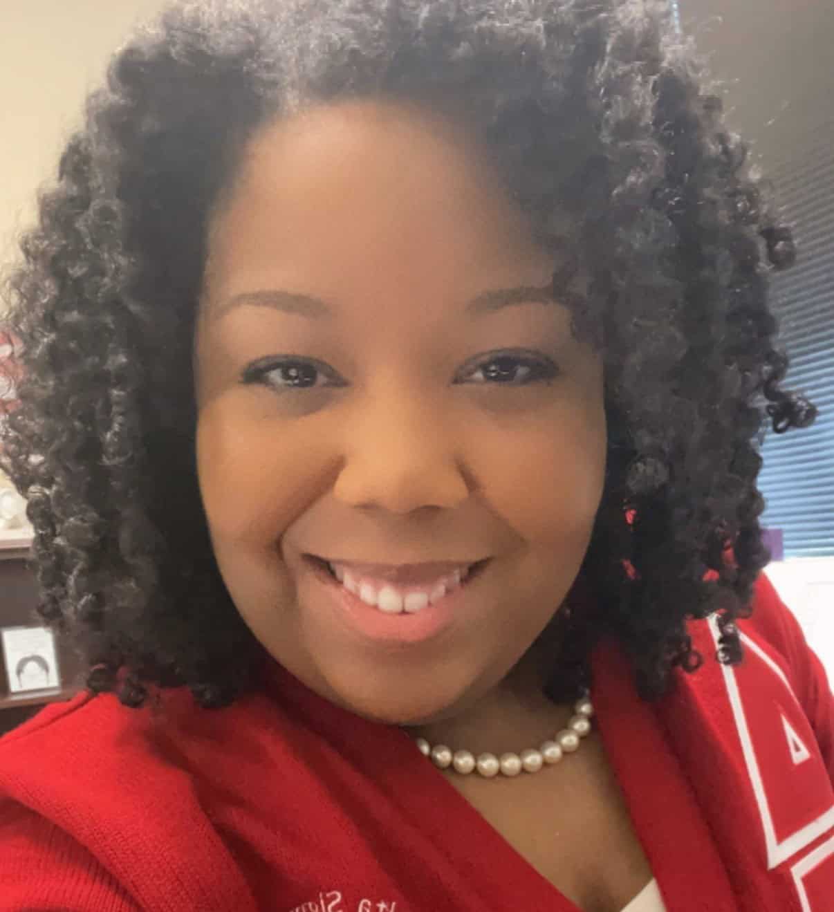 Smiling woman with curly hair wearing a red top and pearl necklace