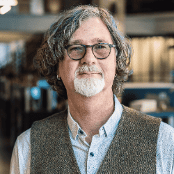 Portrait of a man with glasses and a gray beard wearing a vest and collared shirt