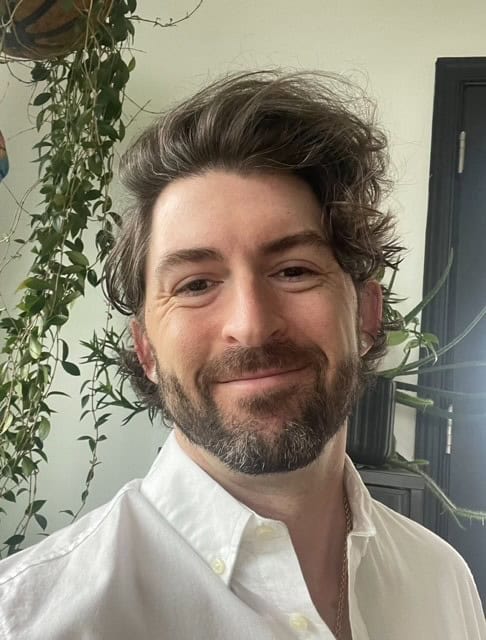 Smiling man with tousled hair and a beard in a white shirt indoors
