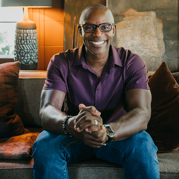 Smiling man in glasses sitting on a couch with his hands clasped
