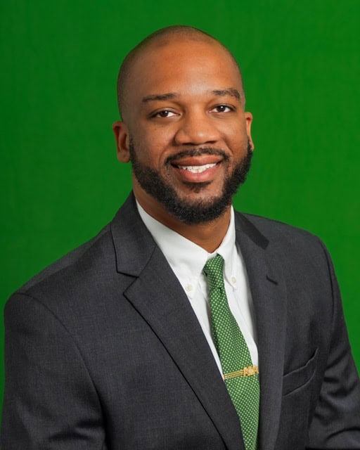 Portrait of a smiling man in a dark suit and green tie against a green background