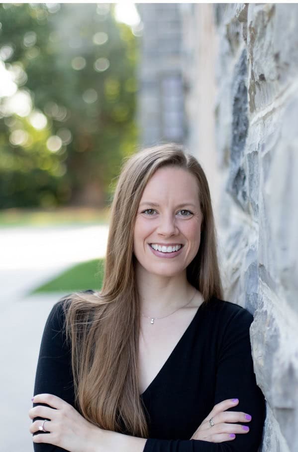 Smiling woman with long hair standing outdoors beside a stone wall