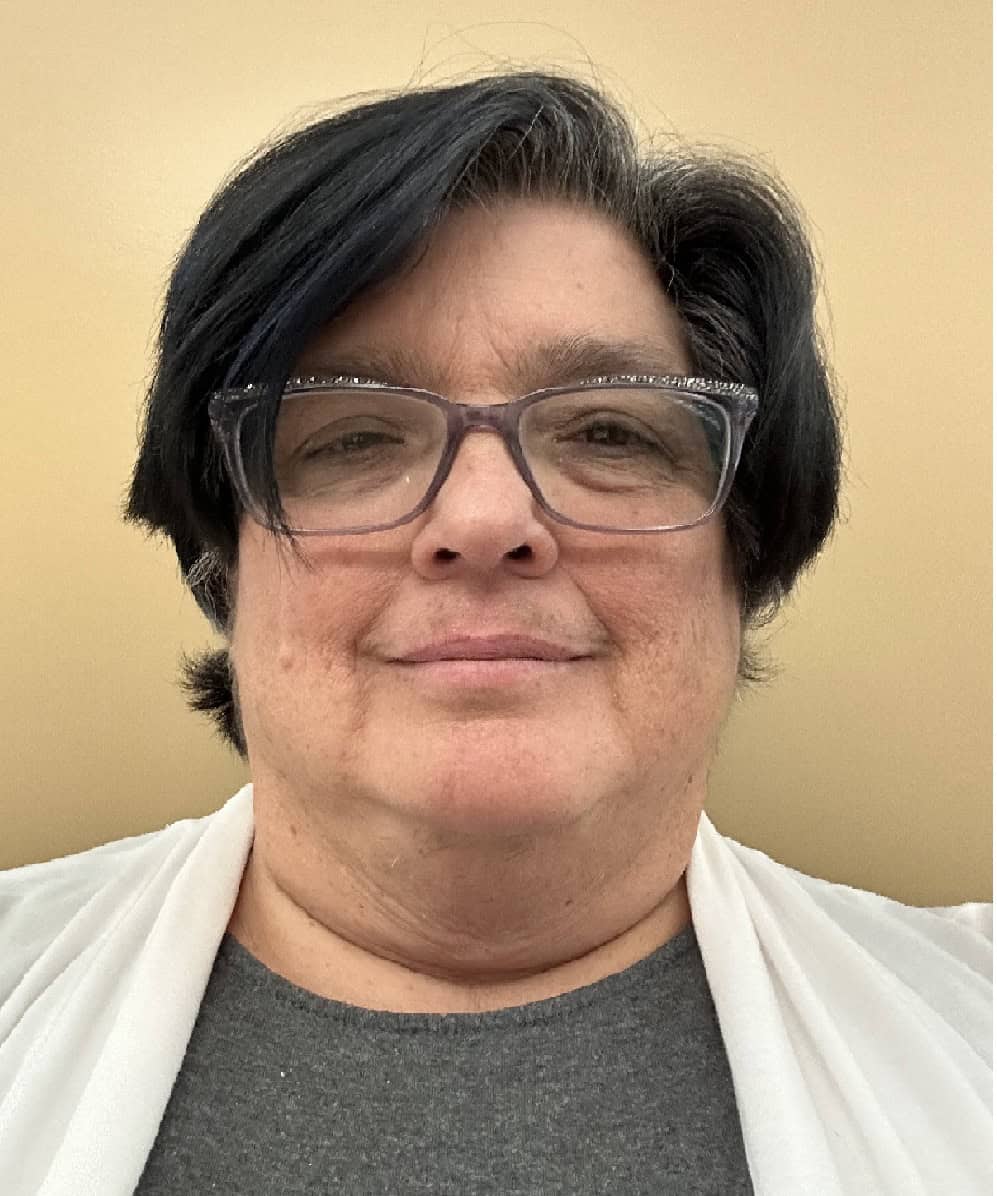 Portrait of a person with short dark hair and glasses, wearing a gray shirt and white cardigan