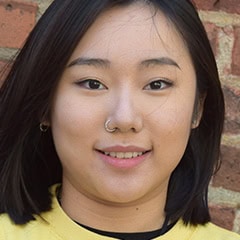 Portrait of a young woman with short dark hair and a nose ring, smiling in front of a brick wall