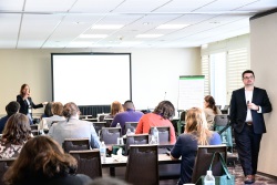 Students sitting in a classroom as a man in a suit jacket walks along the side of the classroom and a female points at the front of the room.