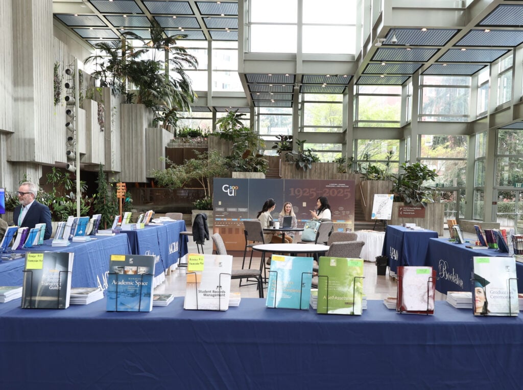 Conference exhibit hall with AACRAO-branded tables of publications and attendees discussing at the event