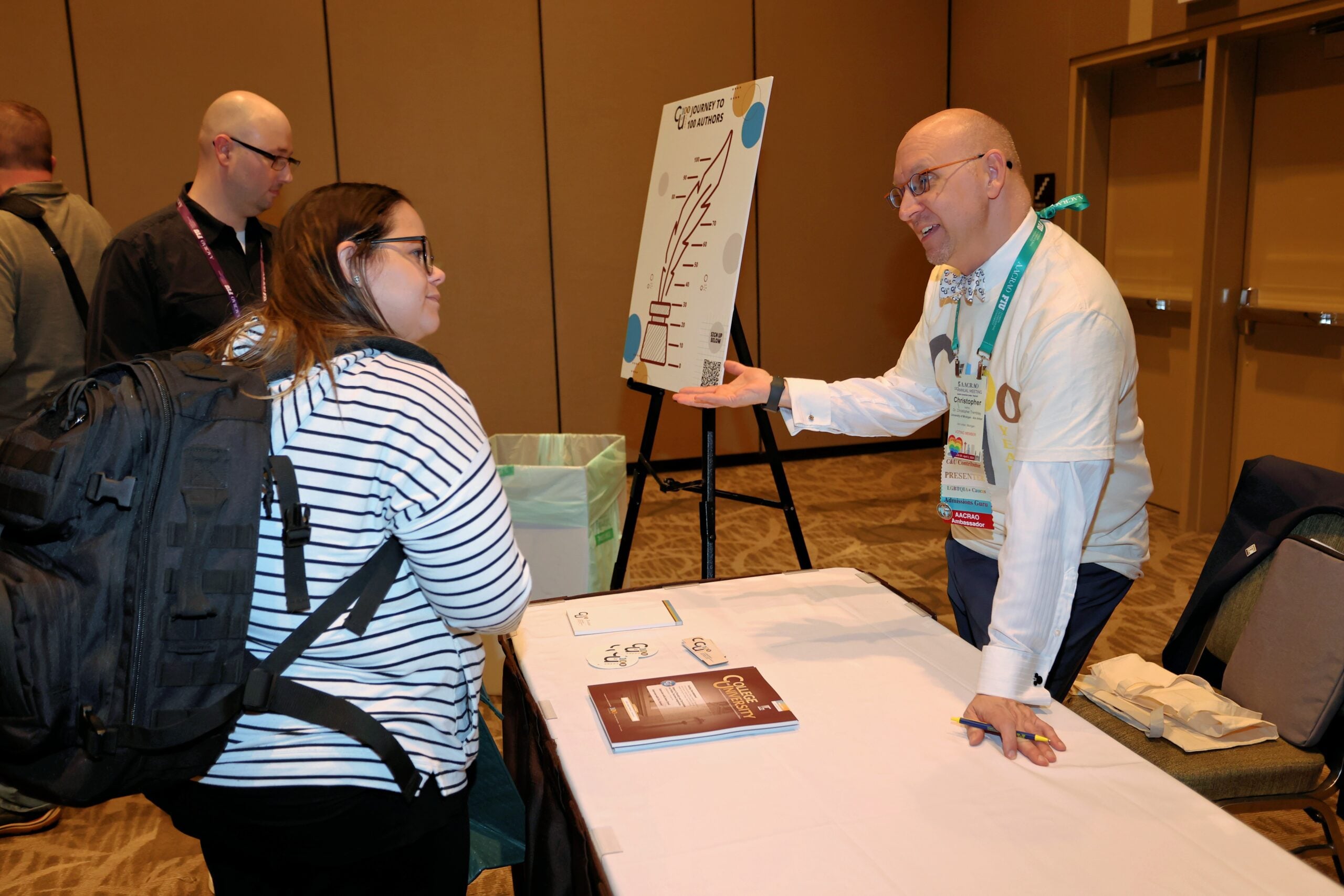 Two attendees discuss a presentation at an AACRAO conference table, with a &ldquo;Journey to 100 Authors&rdquo; poster on an easel