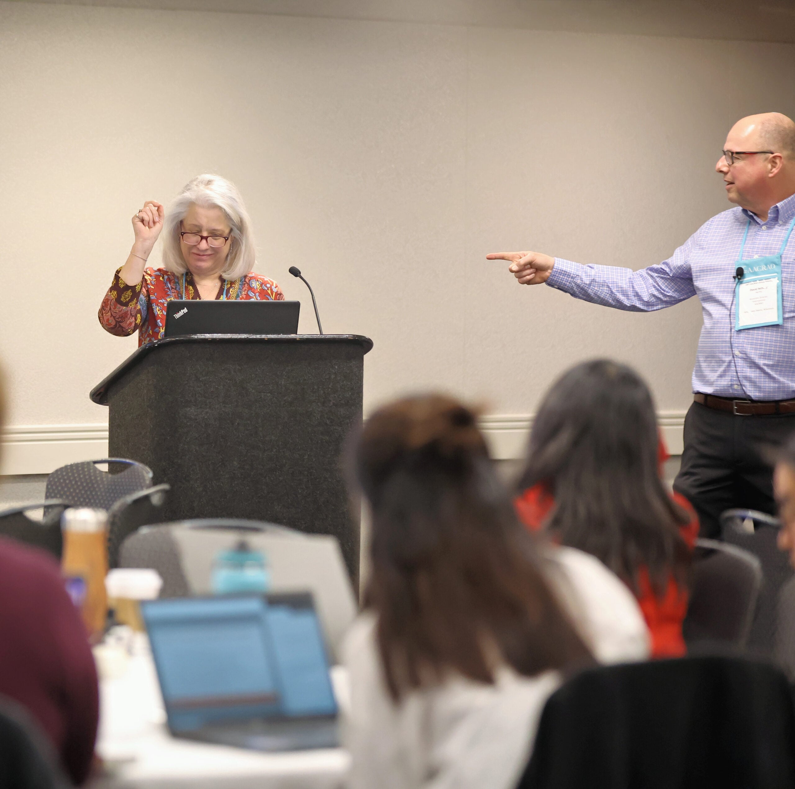 Conference session with a speaker at a podium and another presenter gesturing toward the audience