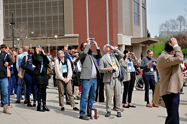 A crowd of people outdoors looking up and taking photos with their phones and cameras