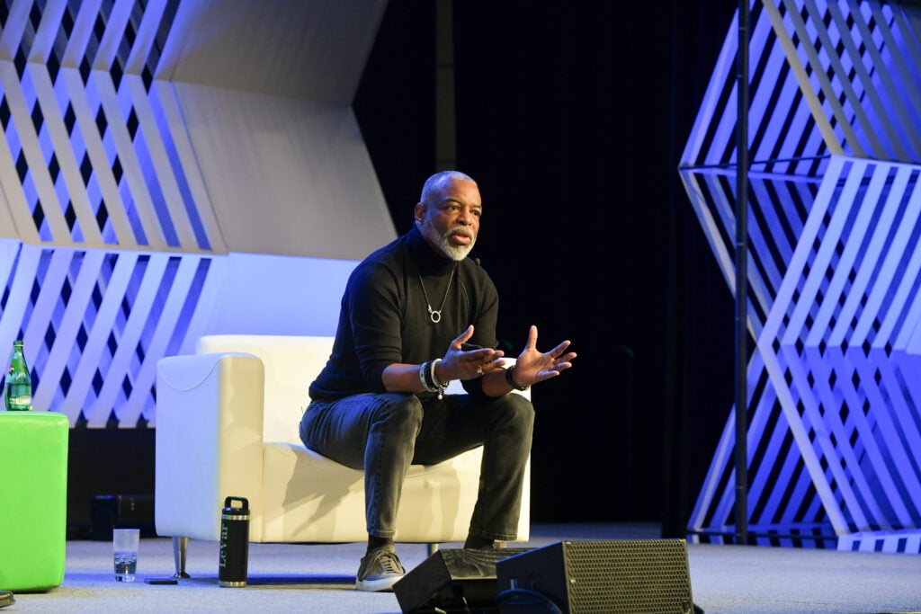 Man speaking onstage at an AACRAO event, gesturing while seated on a panel chair