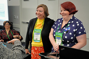 Two women at a conference, smiling and talking while standing near a table with name badges