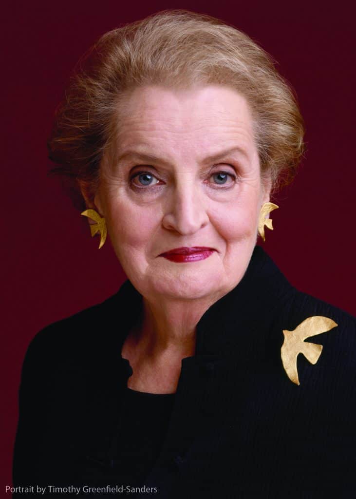 Portrait of a woman with short gray hair wearing gold earrings and a dark top against a maroon background