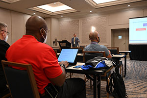 black male wearing a red shirt takes notes on his computer while listening to a presentation