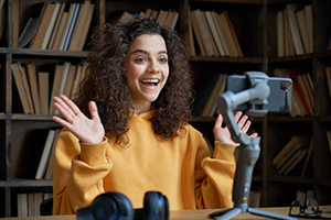 female in orange sweater smiles and waves excitedly while recording herself with her phone