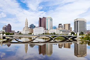 Downtown city skyline reflected in a calm river, with buildings and a bridge in the foreground