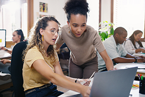 Female teacher helping a female student with something on a laptop.