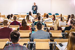 classroom filled with individual desks with seated students facing a teacher standing at the front of the room