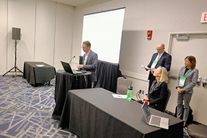 People standing and presenting at a conference meeting room with tables and a screen backdrop