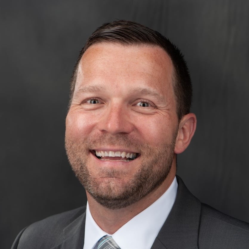 Professional headshot of a smiling man in a suit and tie against a dark background