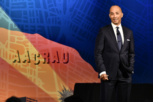 male speaker in a black suit and tie stands in front of a color screen with "AACRAO" displayed