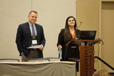 A male and female, both dressed in suits, standing at a podium as though they are presenting.