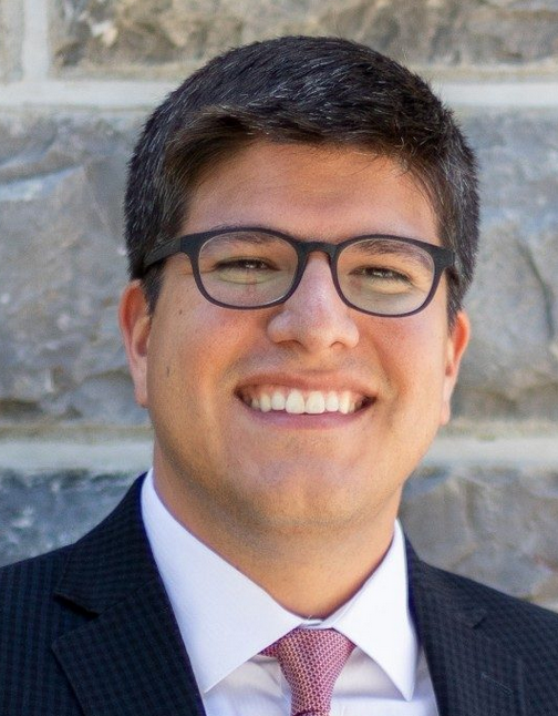 Headshot of a smiling man wearing glasses, a suit, and a pink tie