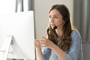 female wearing a blue top sitting in front of a computer monitor while wearing a headset with mic