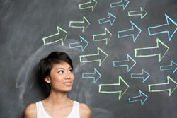 Female looking off to her left as she stands in front of a blackboard with dozens of arrows also pointing to her left.