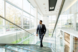 Male wearing a suit walking up a glass staircase in a modern white-walled building.